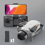 A foldable drone in light grey and black is shown with a matching remote controller and a smartphone mounted on top, plus a small aircraft battery pack and an 'x1' label on a clean gray background.
